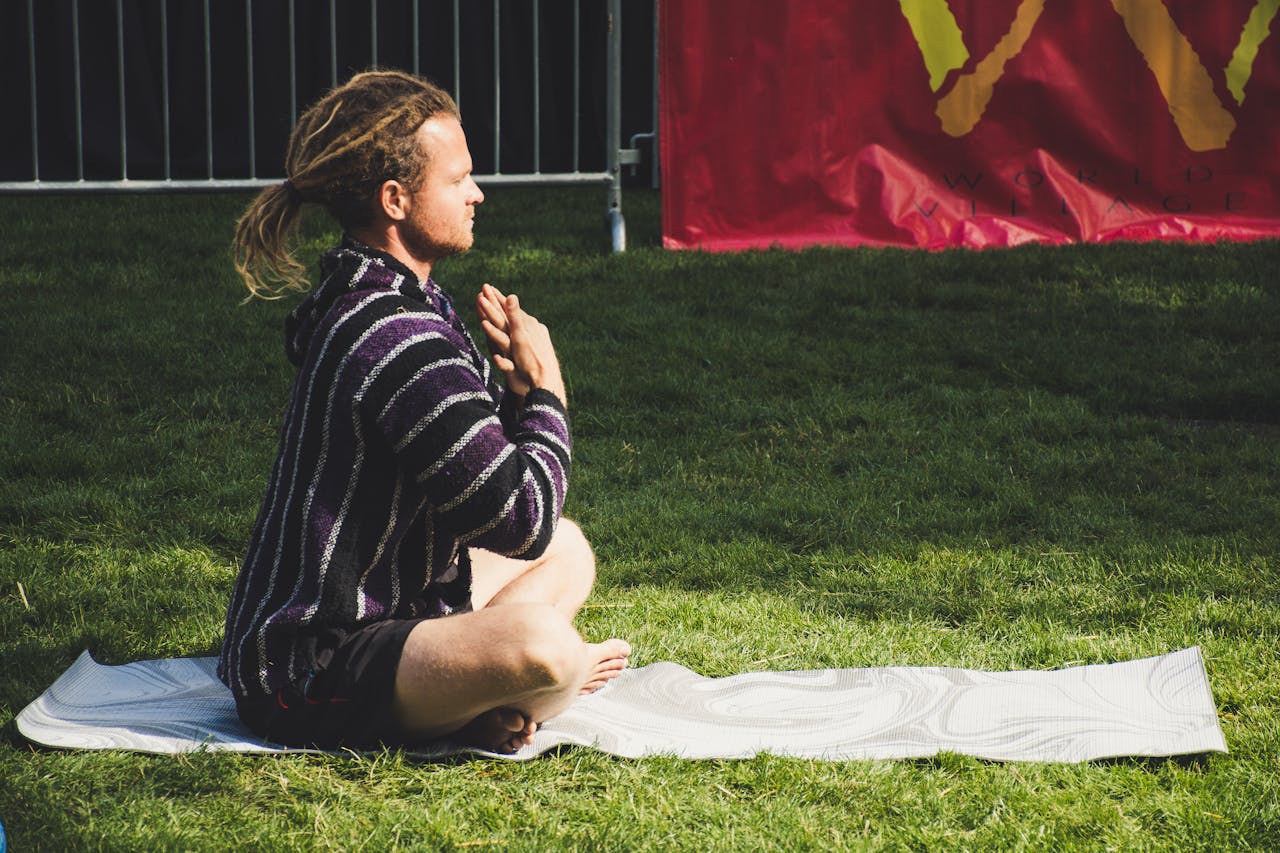 A man meditates in a seated yoga pose on a mat outdoors, enjoying the sunny day.