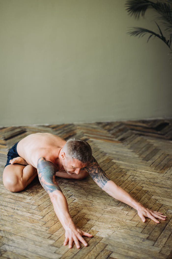 A tattooed man engaging in yoga stretches on a wooden floor indoors.